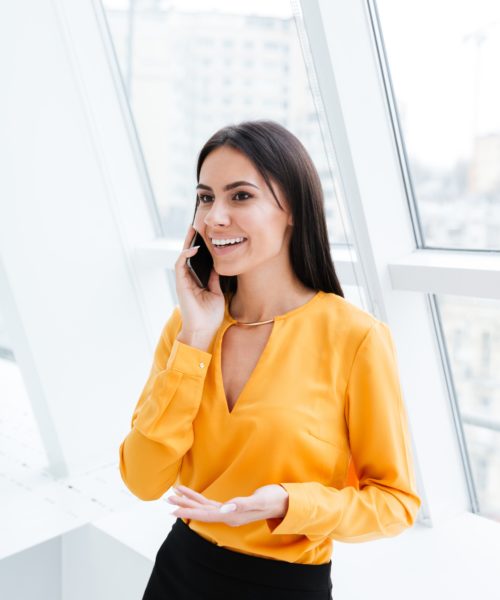 Business woman in orange shirt standing near the window and talking at phone in office